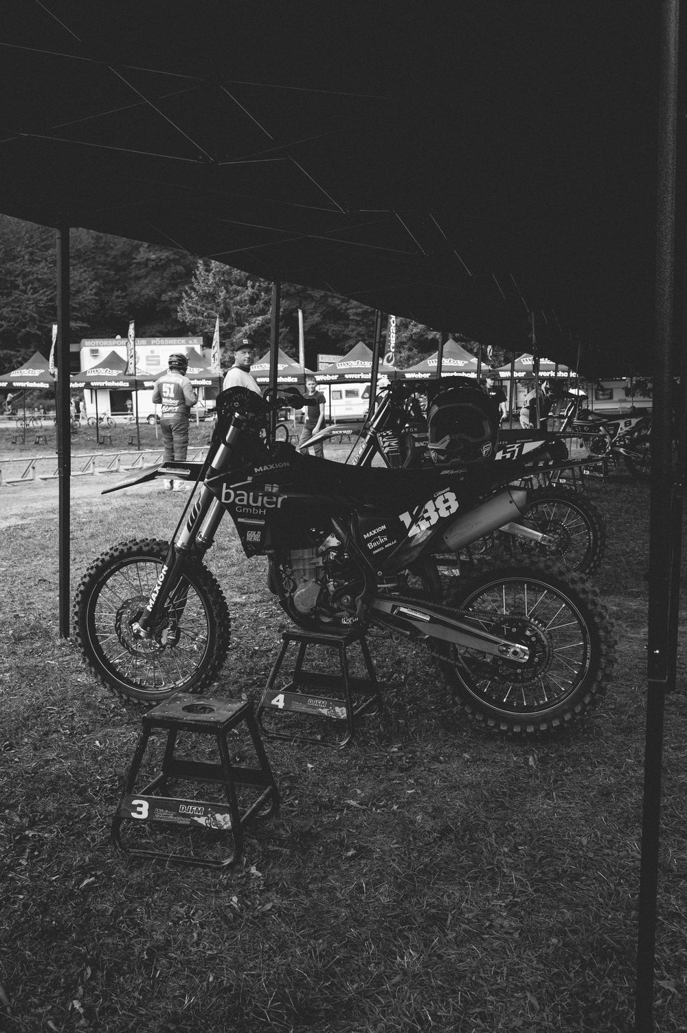 Black and white photo of dirt bikes parked under a tent canopy at a motocross racing event.