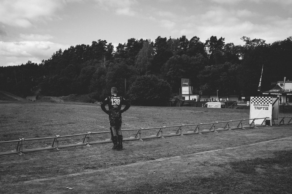 A motocross rider in black gear stands by a racetrack fence with trees and a checkered flag structure in the background.