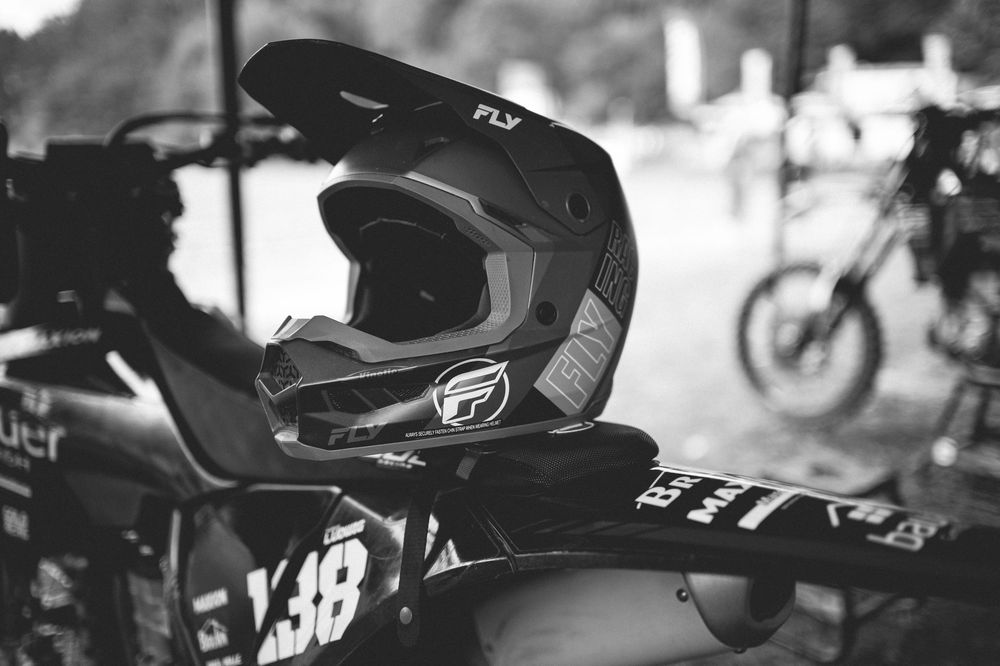 Black and white photo of a Fly Racing motocross helmet resting on a dirt bike with number 38, outdoors.