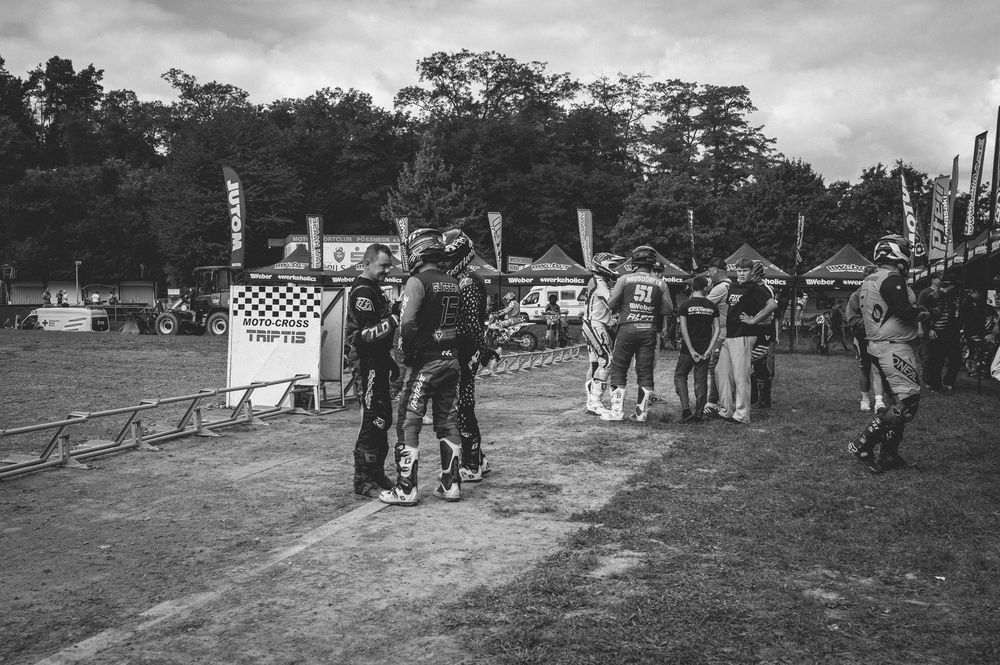 Black and white photo of motocross riders and spectators gathered at an outdoor dirt bike event near a wooded area.
