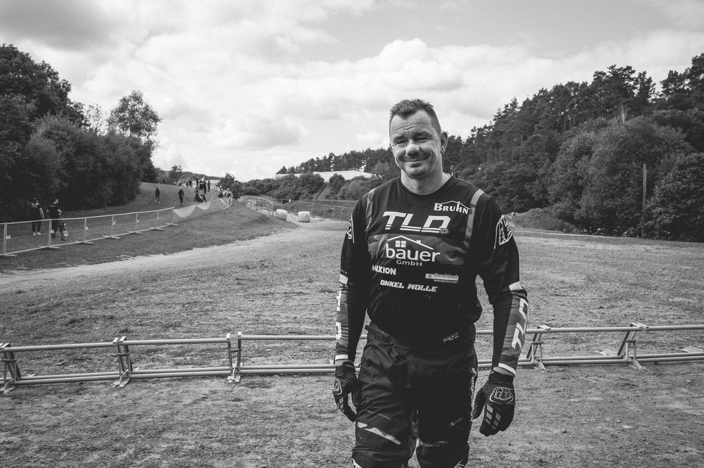 Man in TLO Bauer racing gear smiling at motocross track in black and white photo.