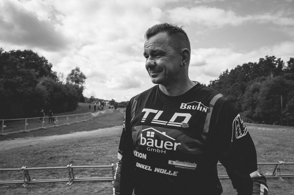 Man in TLD Bauer motocross jersey smiling outdoors at a dirt track race event in black and white.