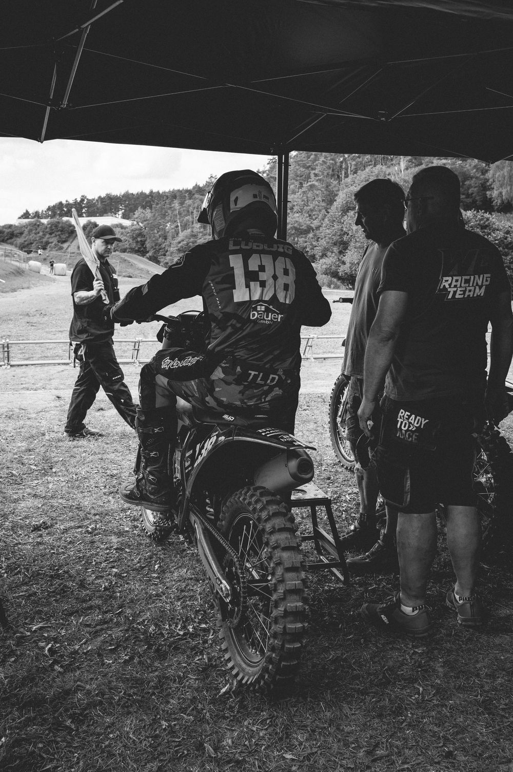 Black and white photo of motocross rider #138 with their bike under a tent, surrounded by racing team members outdoors.
