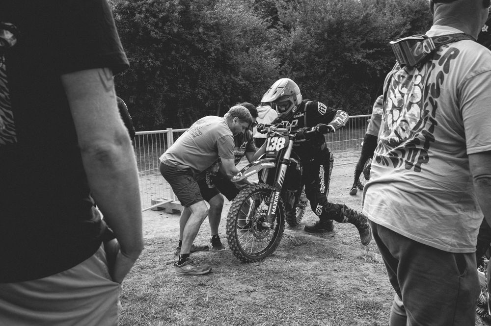 Black and white photo of motocross riders and crew assisting a dirt bike rider numbered 38 at an outdoor racing event.