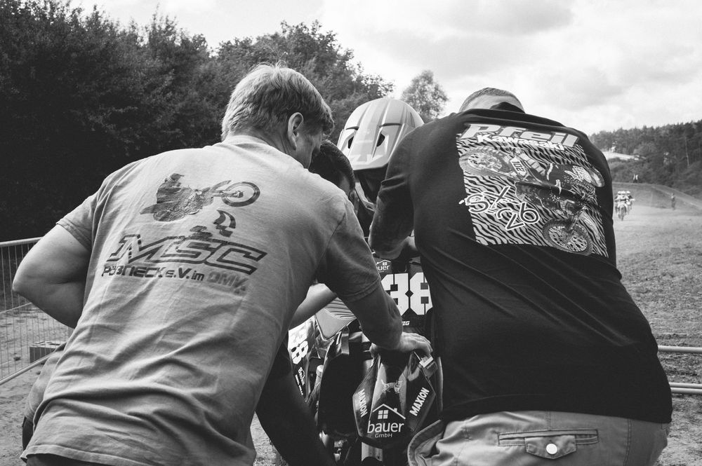 Black and white photo of two people in motorsport gear examining a dirt bike at an outdoor racing event.