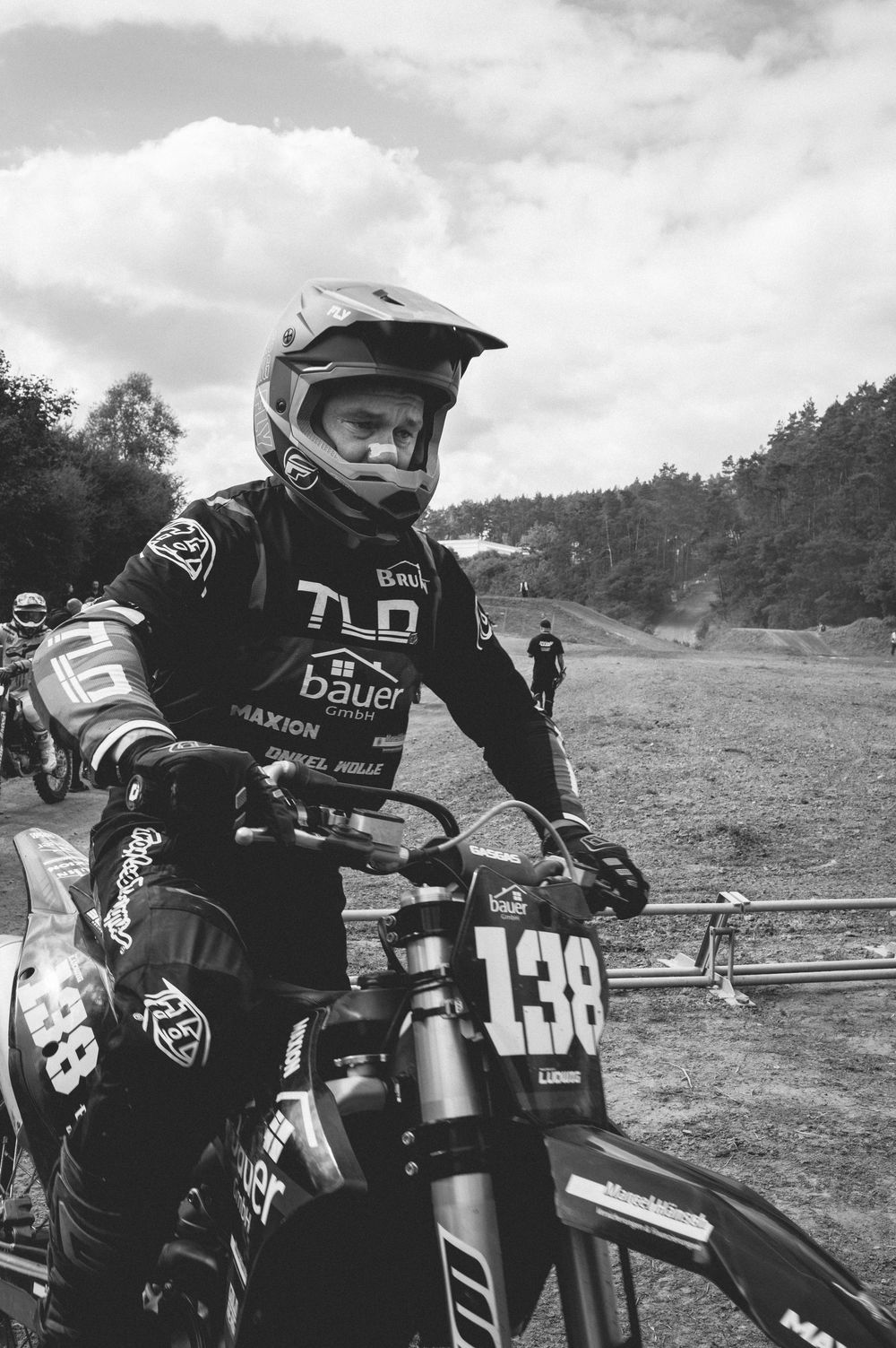 Motocross rider #138 in TLD gear and helmet sits on dirt bike at outdoor race event, black and white photo.