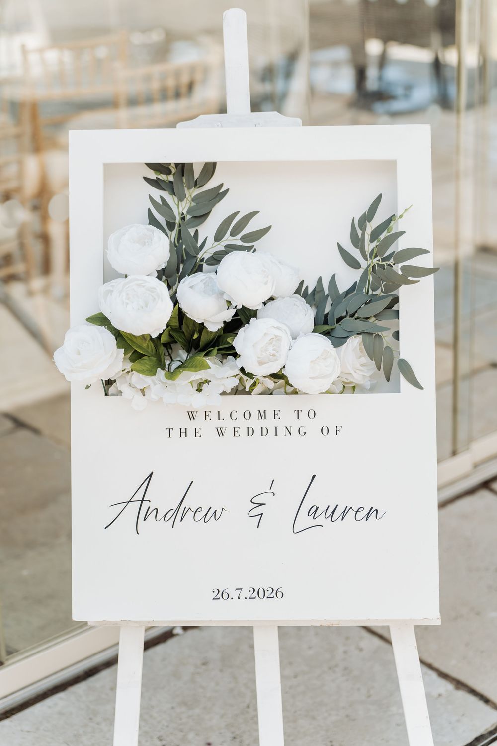 Elegant wedding welcome sign on easel with white roses, greenery, and script text for Andrew & Lauren, dated 26.7.2026.