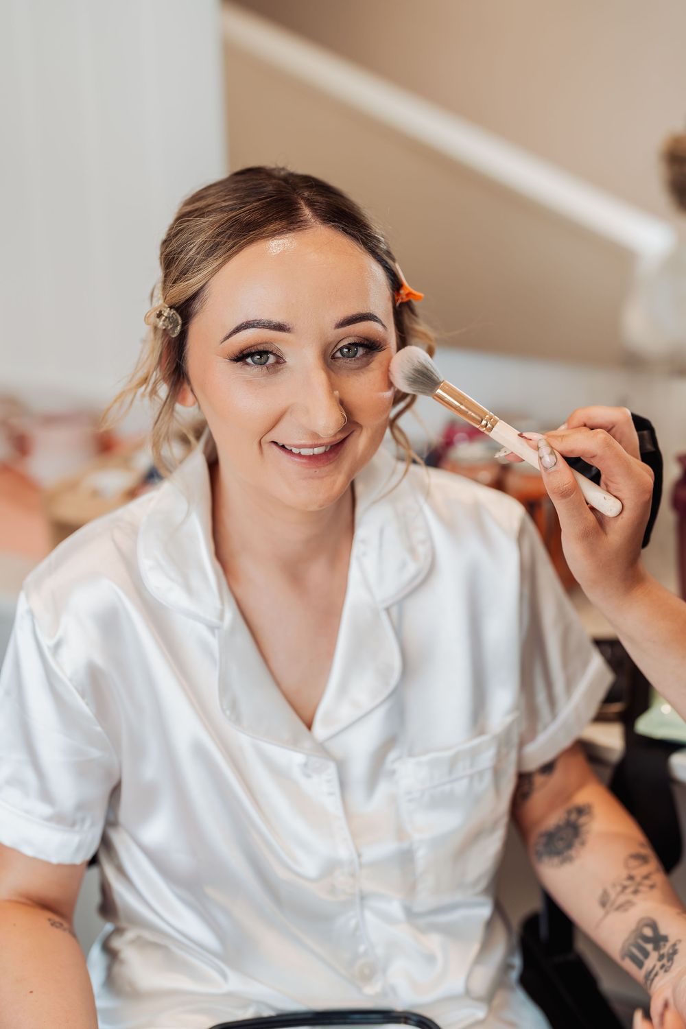 Bride in white satin robe smiling while makeup artist applies blush with a brush during wedding preparation.