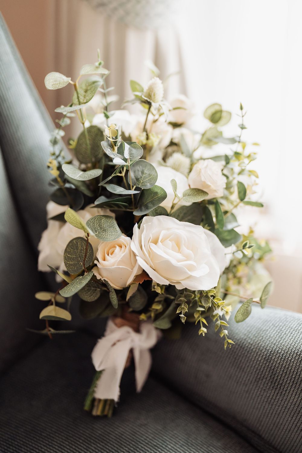 Elegant bridal bouquet of white roses and eucalyptus tied with blush ribbon resting on a gray chair.