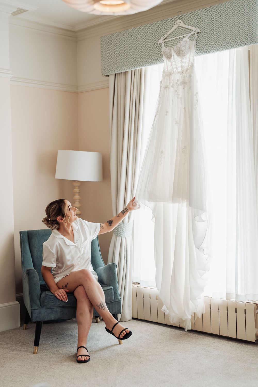 Bride in white robe sits in teal chair reaching toward her wedding dress hanging by a bright window.