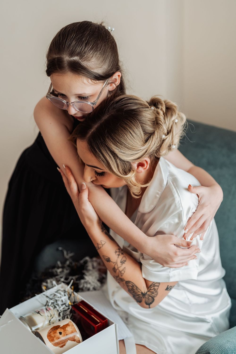 A bride in white robe shares a tender hug with a young girl wearing glasses during wedding preparations.