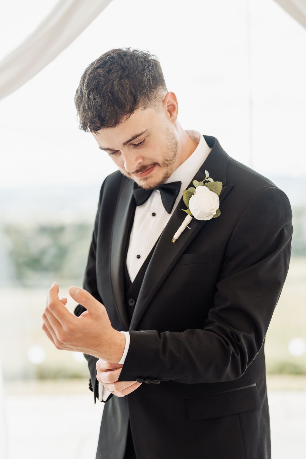 Groom in black tuxedo with white boutonniere adjusting cufflinks before wedding ceremony.