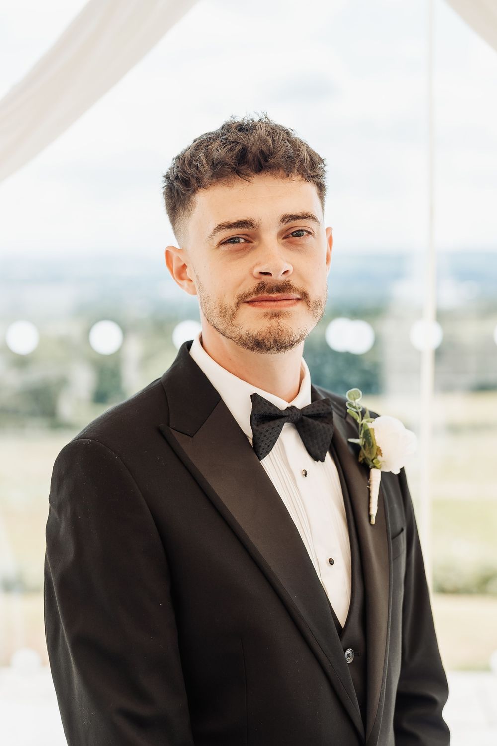 A dapper groom in a black tuxedo with bow tie and boutonniere smiles confidently at a bright outdoor wedding venue.