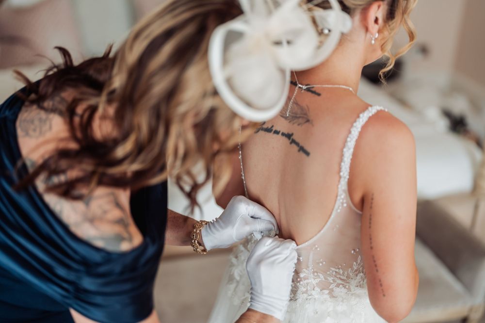 Bridesmaid in blue dress fastening back of bride's lace wedding gown, bride has back tattoo.
