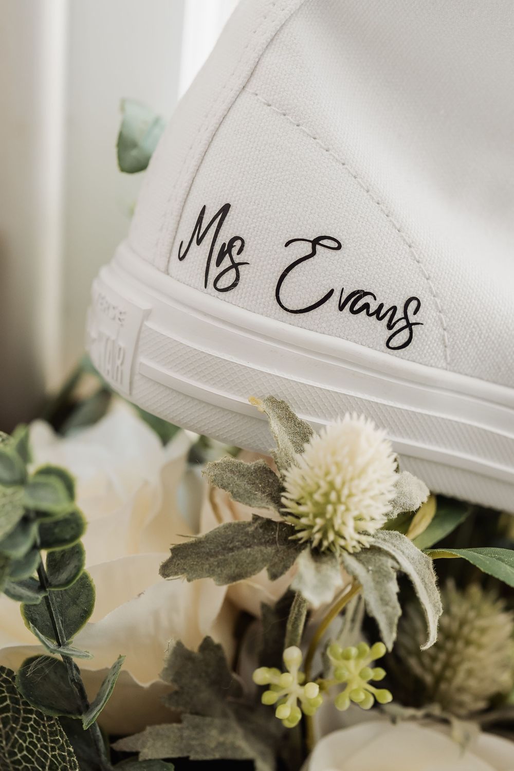 White bridal sneaker with Mrs Evans custom script lettering, surrounded by elegant white wedding bouquet flowers.