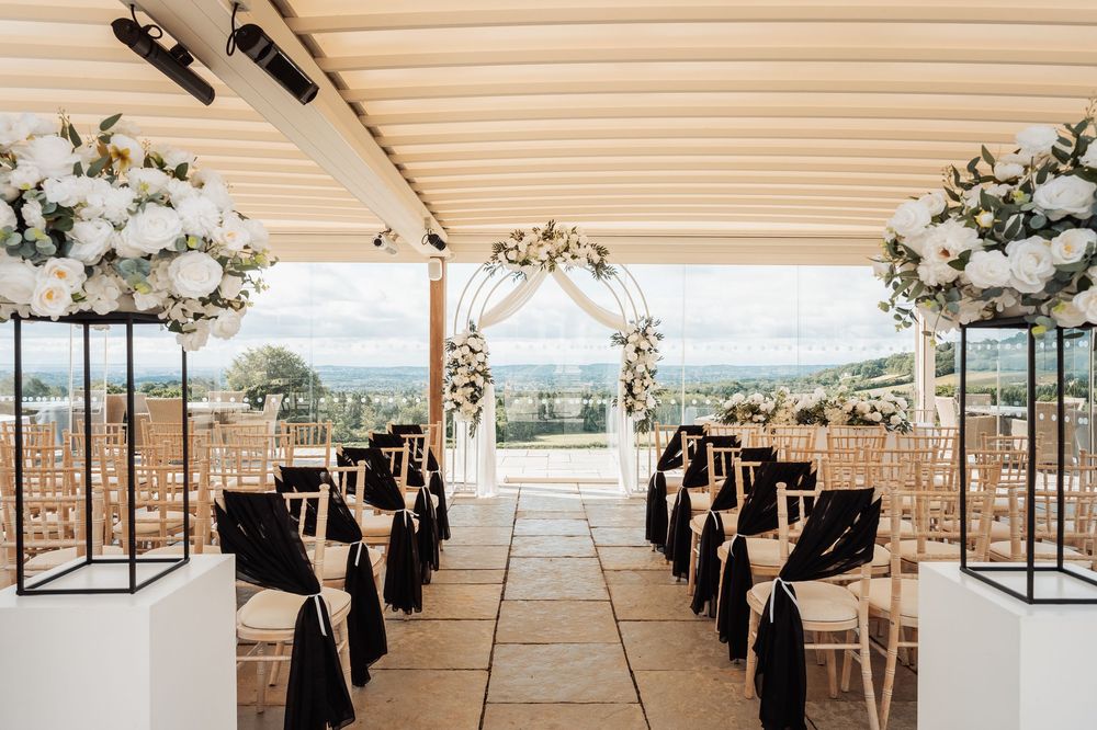 Elegant outdoor wedding ceremony setup with black and white floral arch, chiavari chairs, and panoramic countryside views.