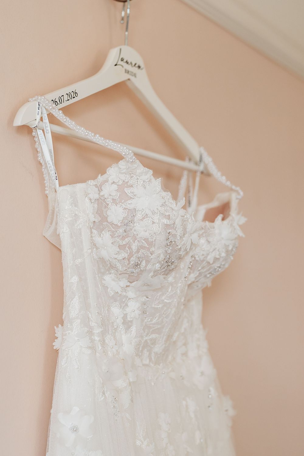 White lace wedding dress hanging on personalized bridal hanger with date 26.07.2026 against blush pink wall.