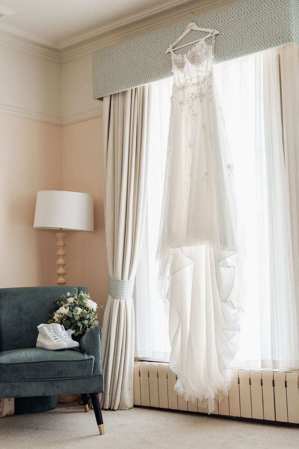 White lace wedding dress hanging in window with bridal bouquet and white sneakers on teal armchair in elegant room.