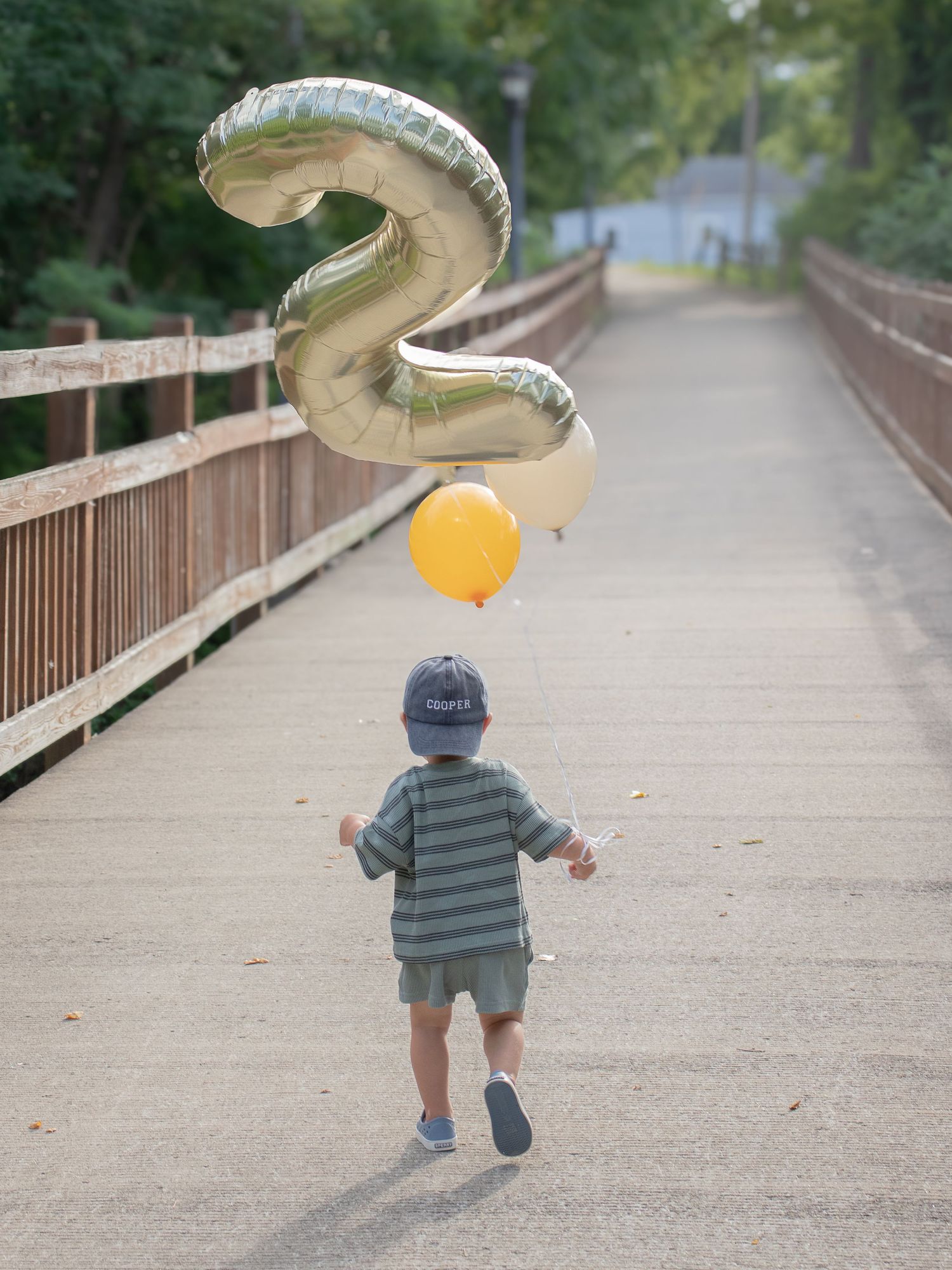 Toddler walking away on wooden bridge holding gold number two and yellow birthday balloons on a tree-lined path.