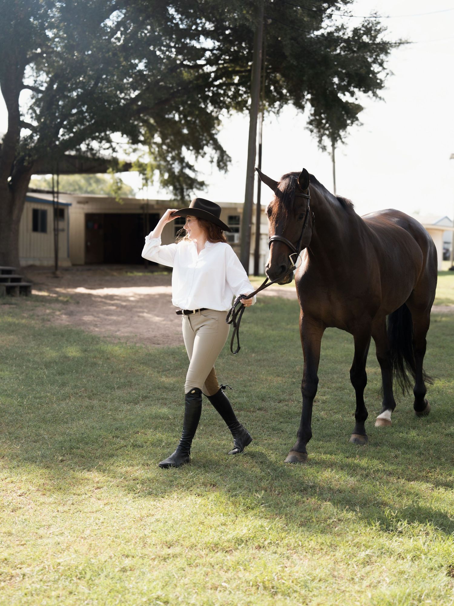 A woman in equestrian attire walks a dark brown horse on a sunny day at a ranch.