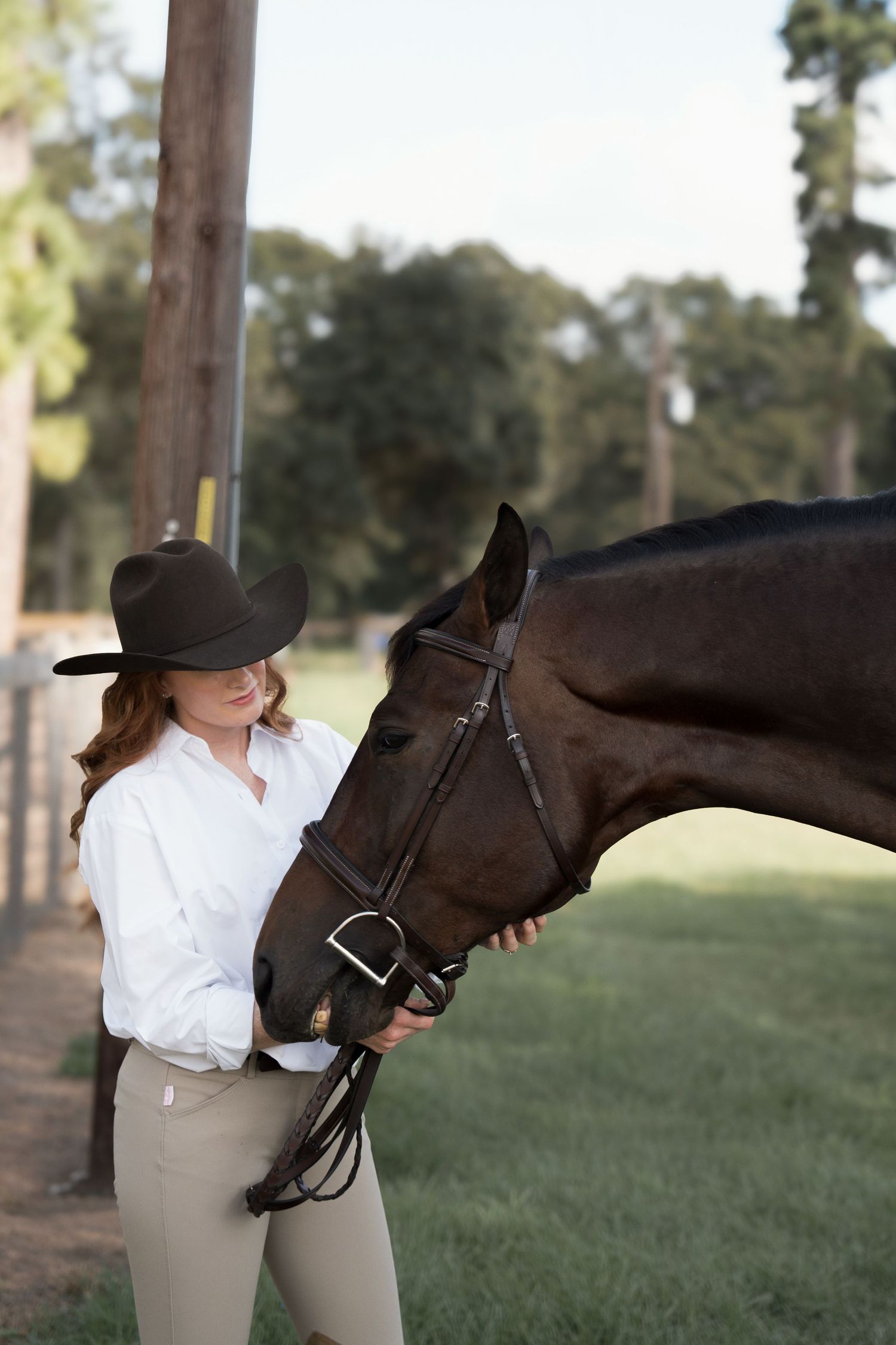 Woman in cowboy hat and white shirt holds bridle of dark brown horse outdoors at equestrian facility.