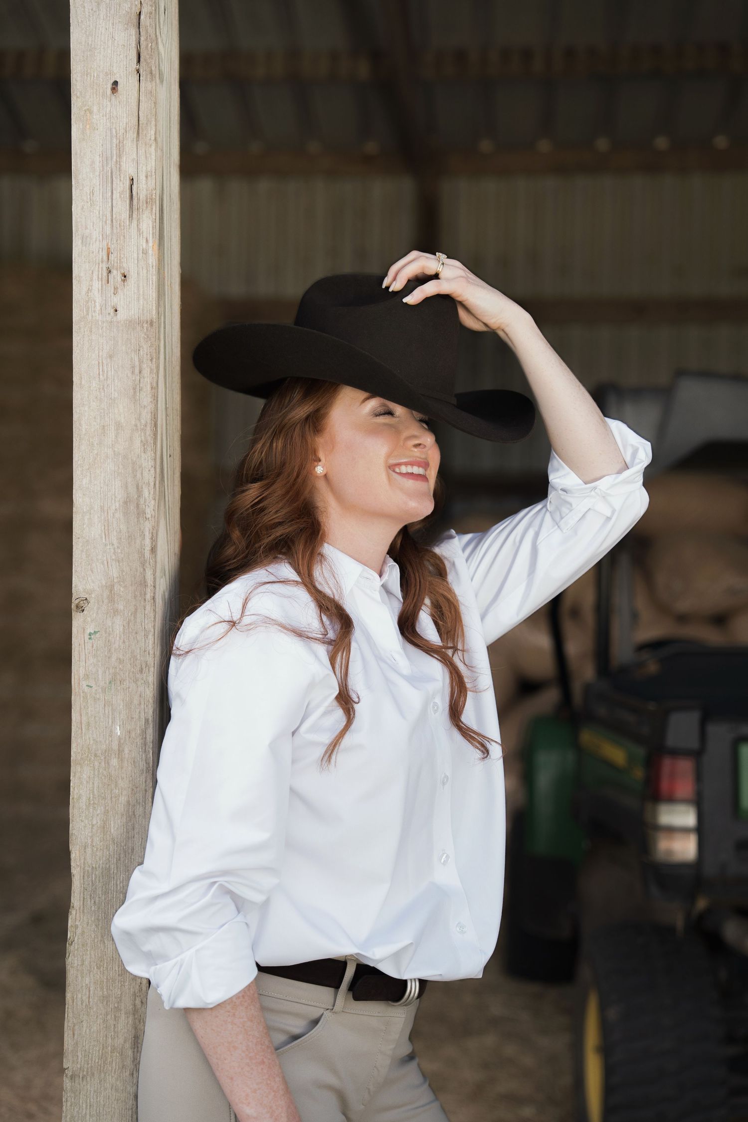 Smiling woman in white shirt and black cowboy hat leaning against a wooden post inside a barn.