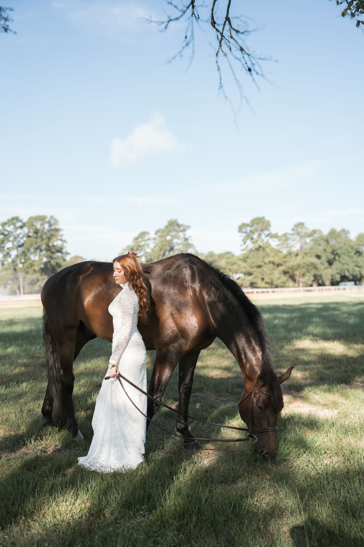 Bride in white lace gown holds reins as brown horse grazes on green pasture under blue sky.