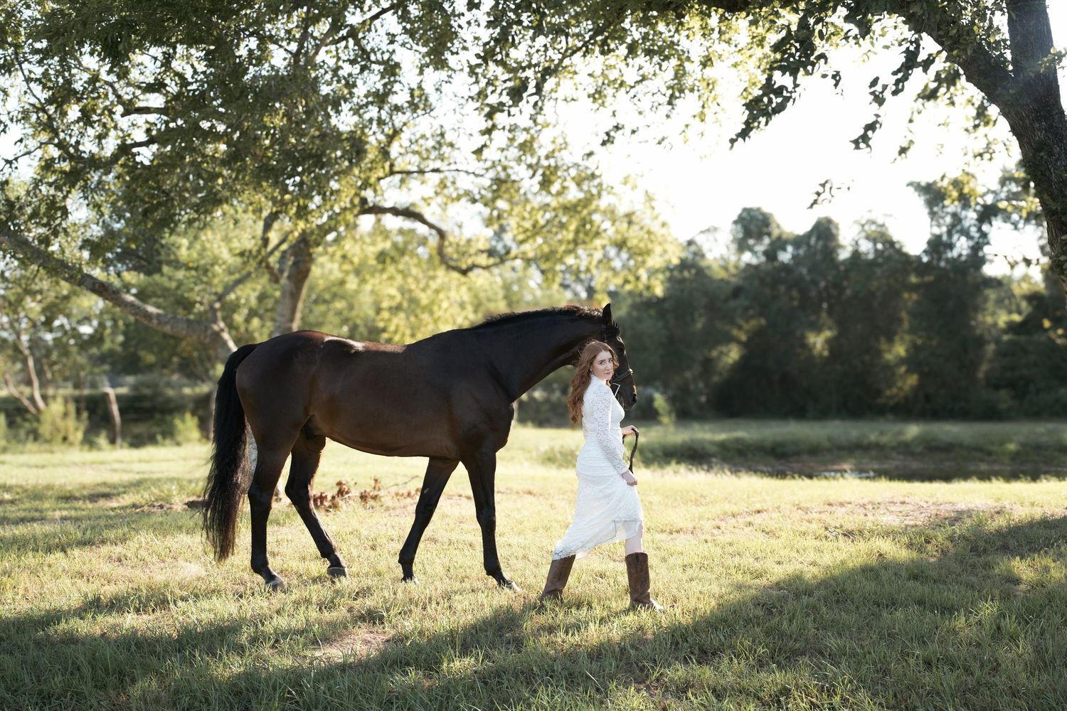 Woman in white dress walking with a dark brown horse in a sunlit green field under large oak trees.