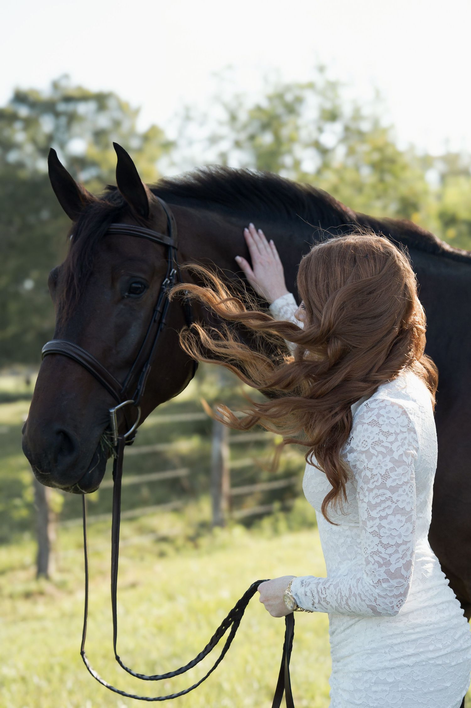 Red-haired bride in white lace dress tenderly touches dark horse face while holding reins outdoors.