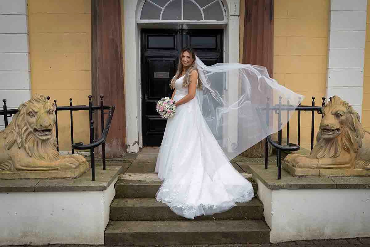 Bride in white wedding gown and flowing veil poses on stone steps at Cockermouth town Hall registry office