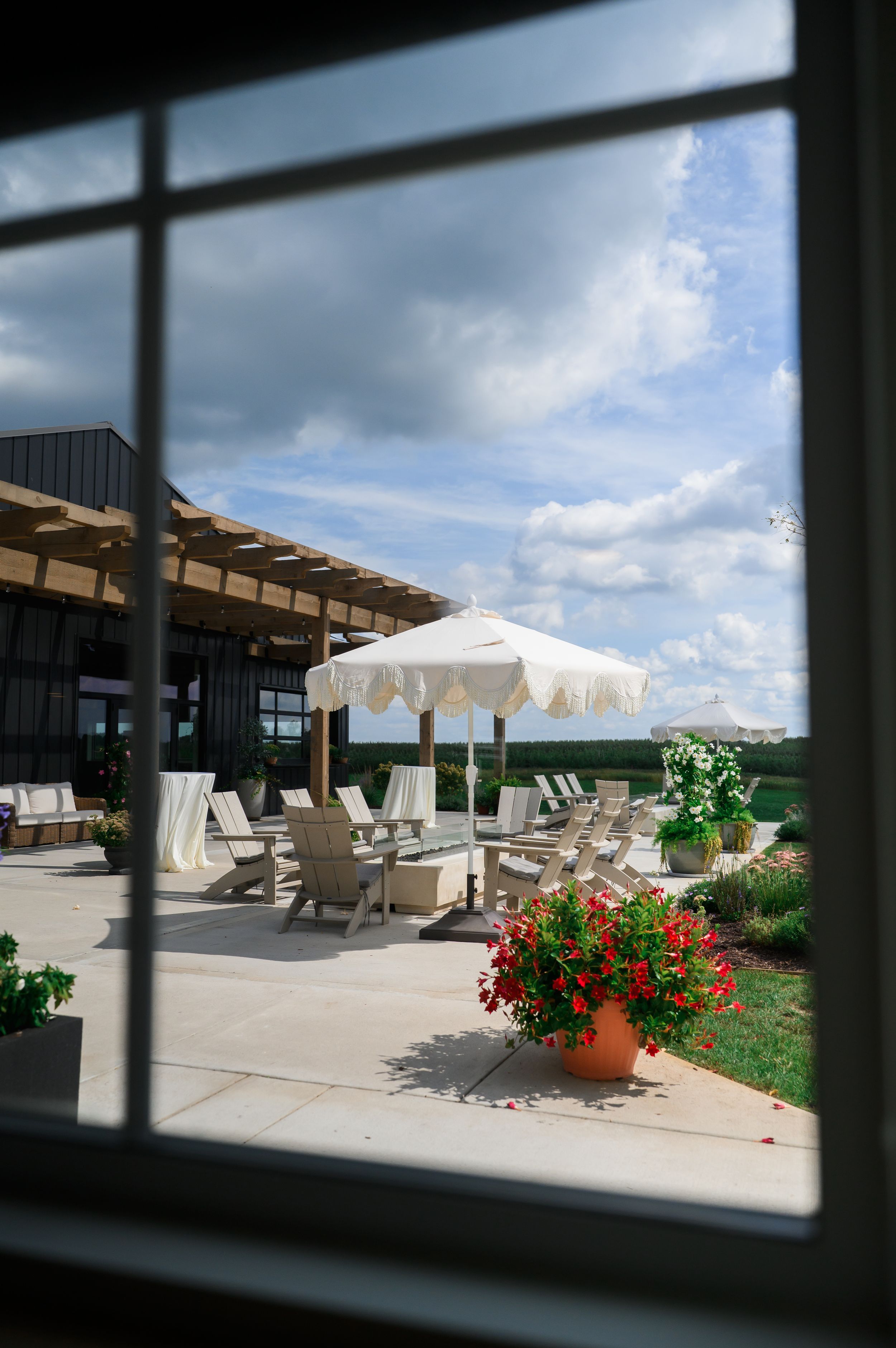 Elegant outdoor patio with white lounge chairs, large umbrella, and vibrant flower pots viewed through a window.