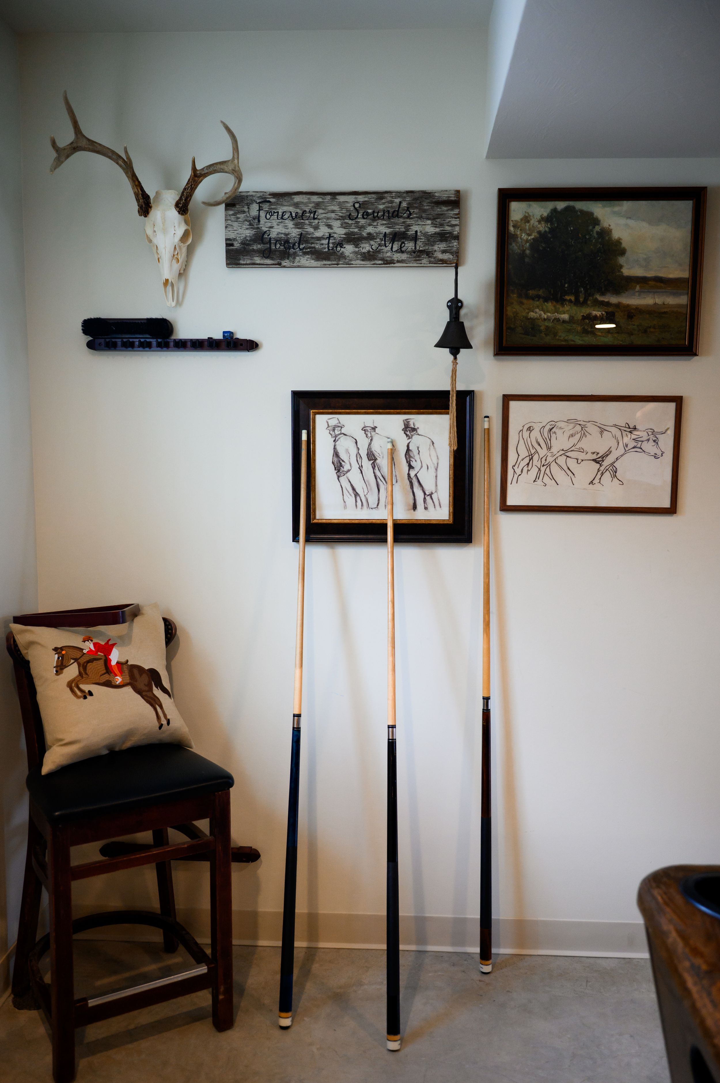 Room corner with pool cues, deer skull, framed equestrian art prints, a barstool with horse pillow, and rustic wall decor.