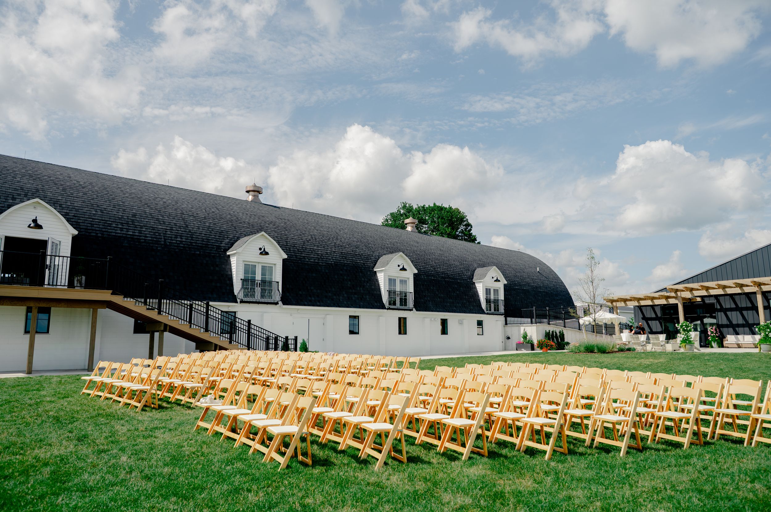 Outdoor wedding ceremony setup with wooden chairs on green lawn beside a white barn venue under a blue sky.