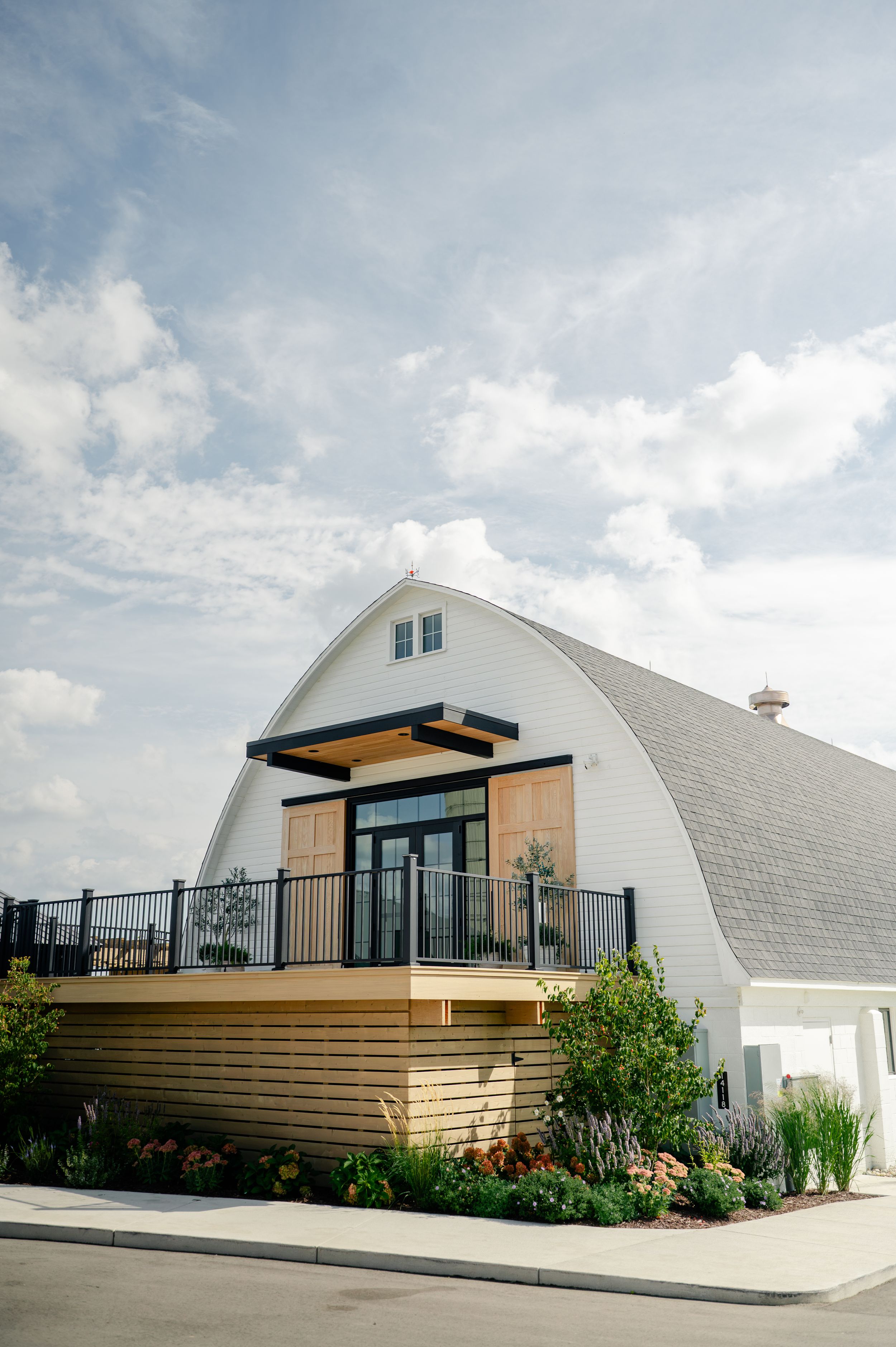 Modern white barn-style building with gambrel roof, wood deck, black railings, and glass doors surrounded by landscaped gardens.