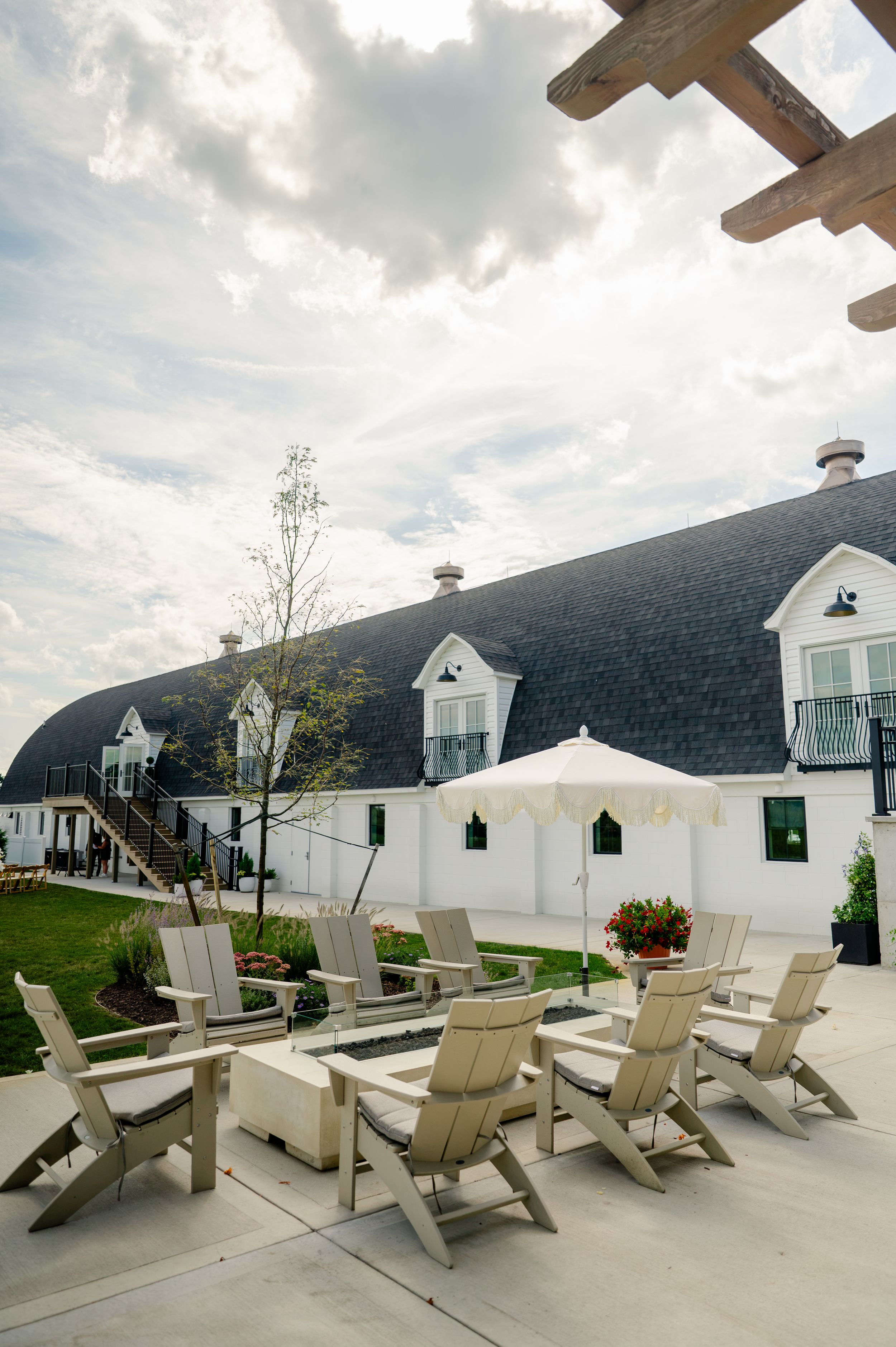 Adirondack chairs arranged around a fire pit patio outside a white barn-style building with dark curved roof under cloudy sky.
