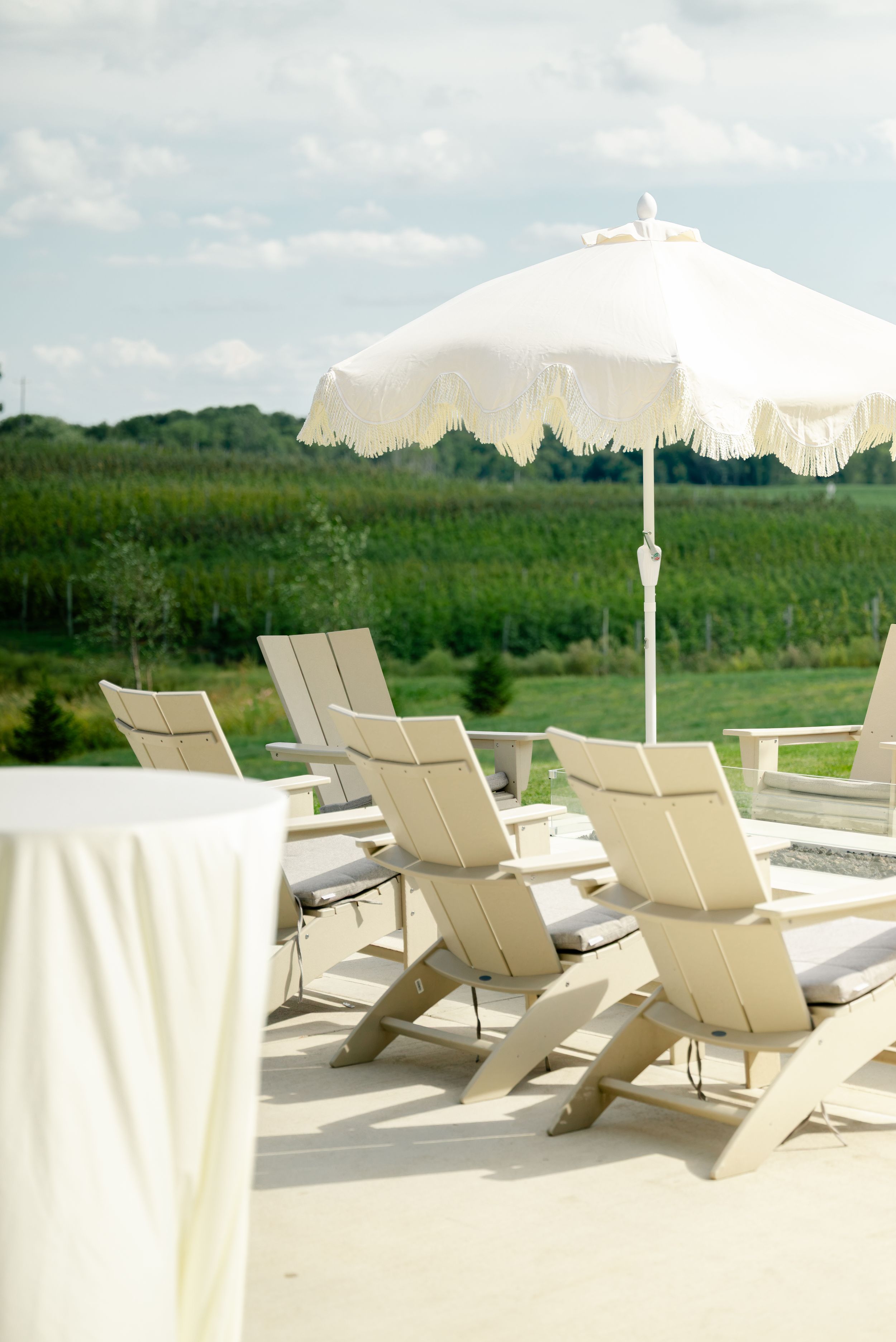 White Adirondack chairs and a fringed umbrella on a patio overlooking lush green vineyard rows under a cloudy sky.