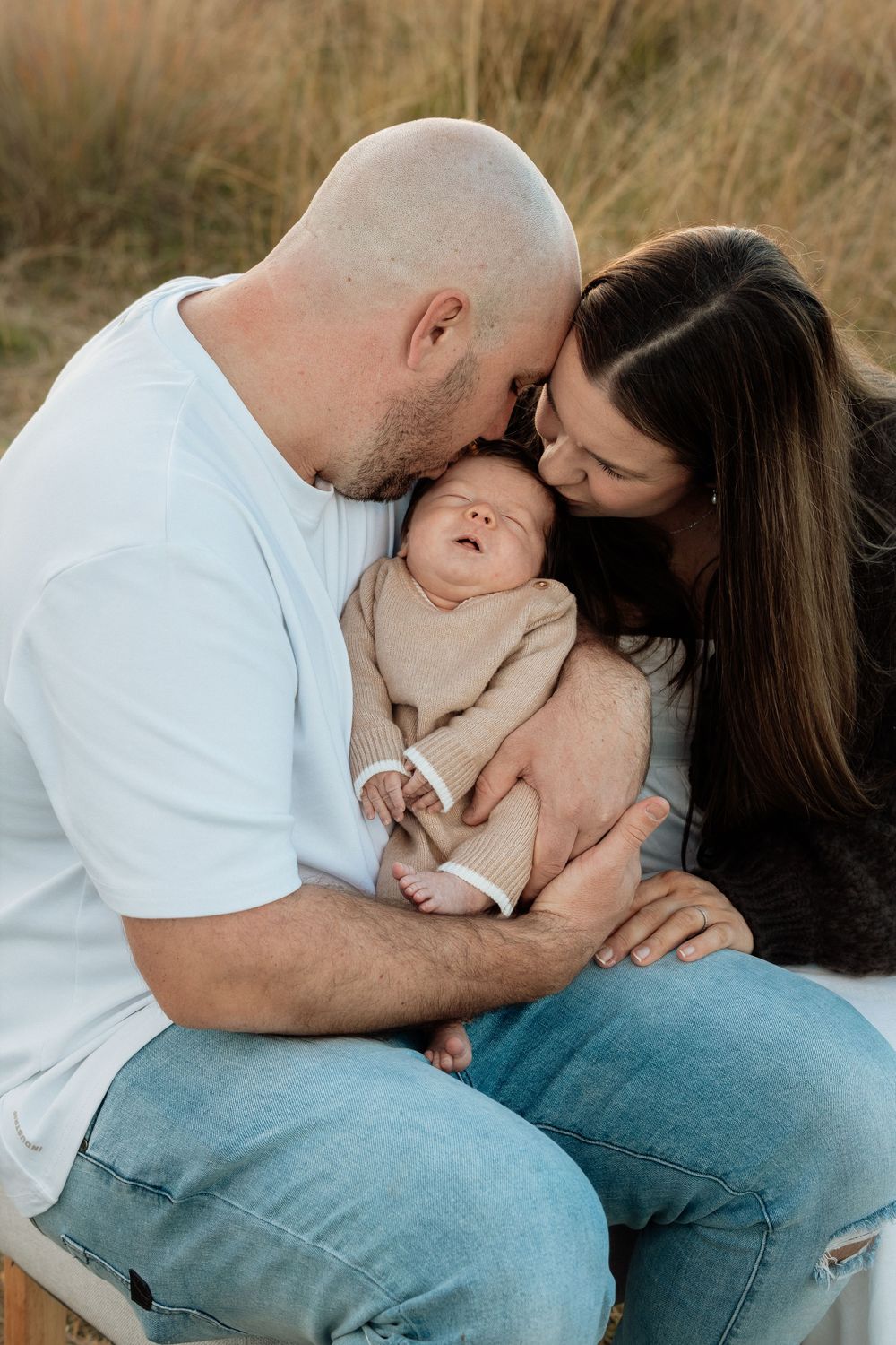 Parents kissing their sleeping newborn baby together in a warm outdoor field setting at Rouse Hill Regional Park.