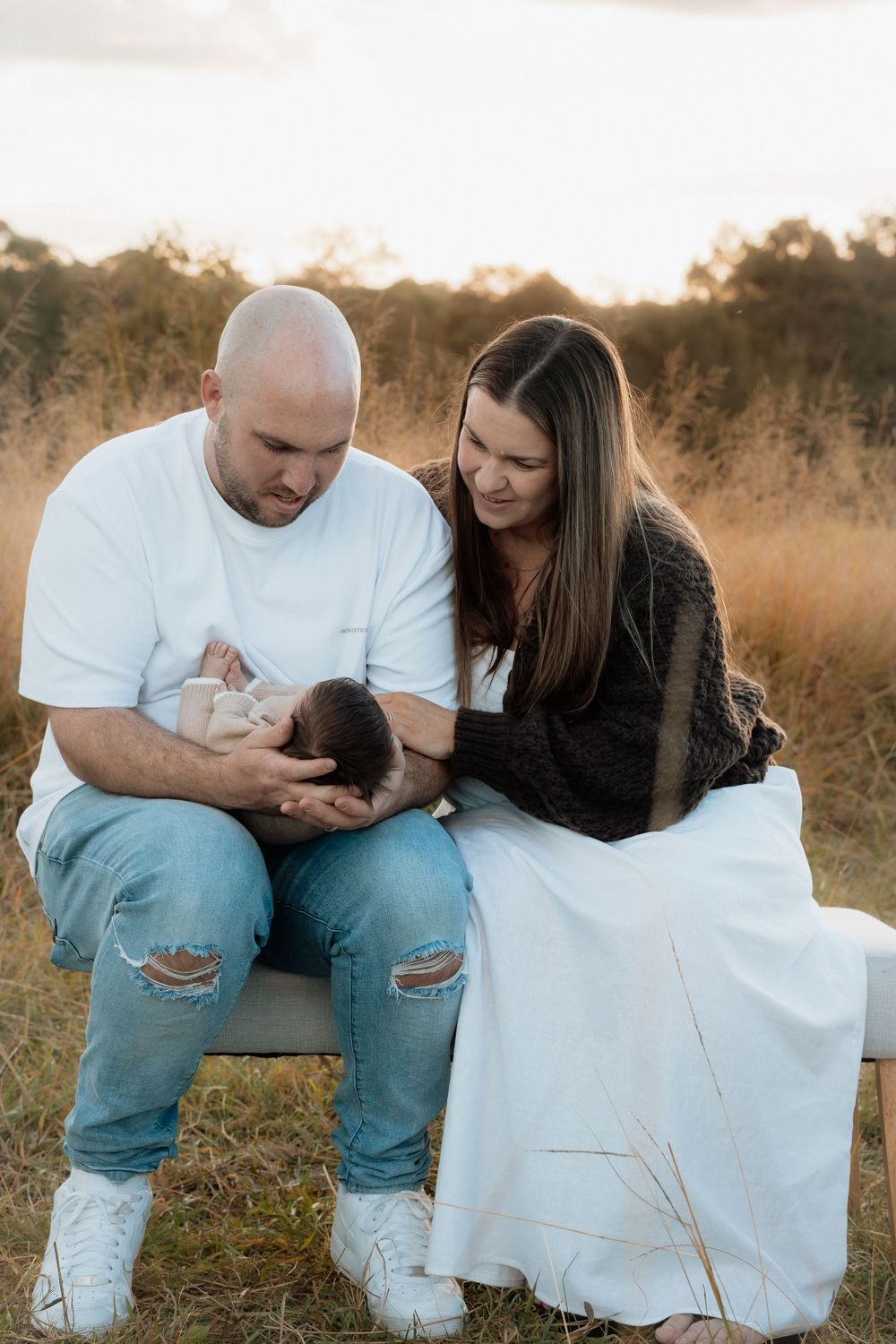 New parents sit outdoors in golden field, father cradling newborn baby while mother lovingly looks on at sunset.