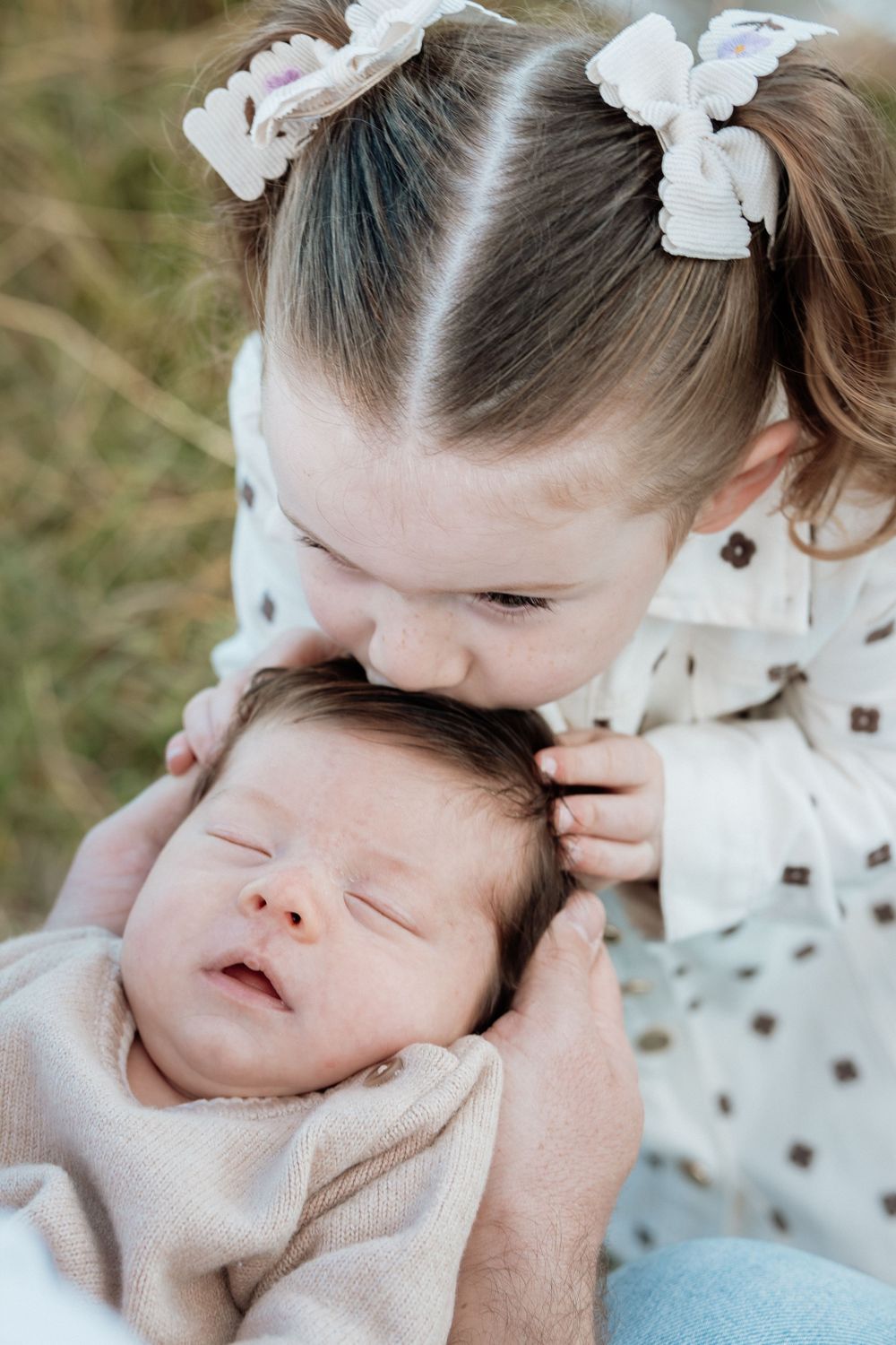 Young girl with bow pigtails gently kisses sleeping newborn baby wrapped in soft beige blanket outdoors.
