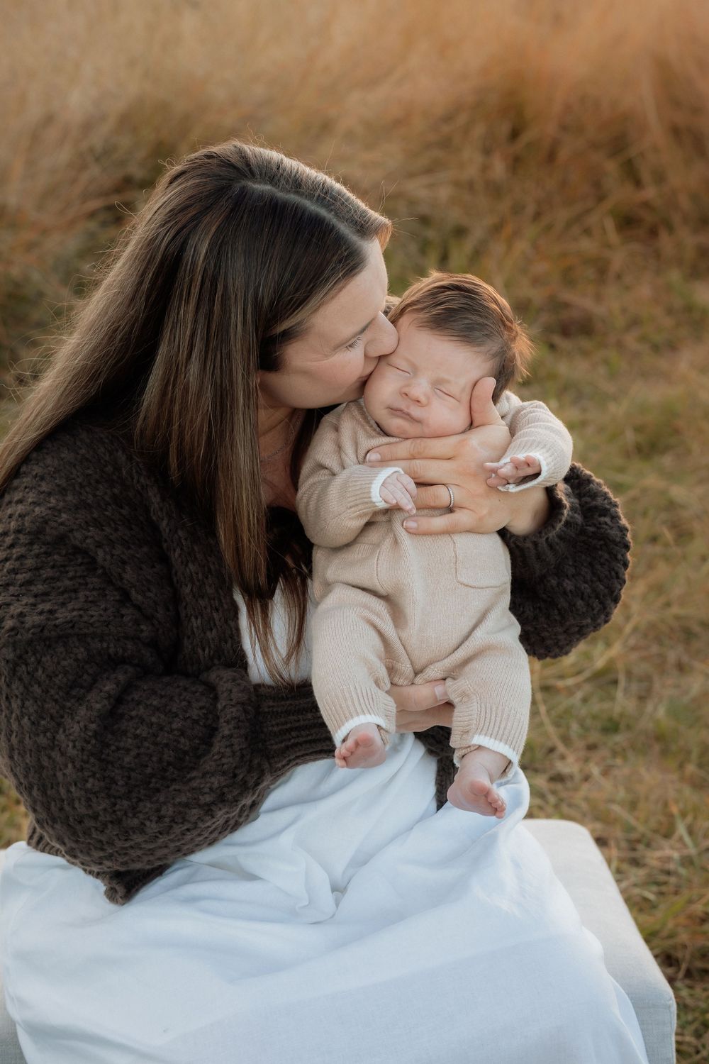 Mother in brown sweater tenderly kissing sleeping newborn baby dressed in beige romper outdoors.