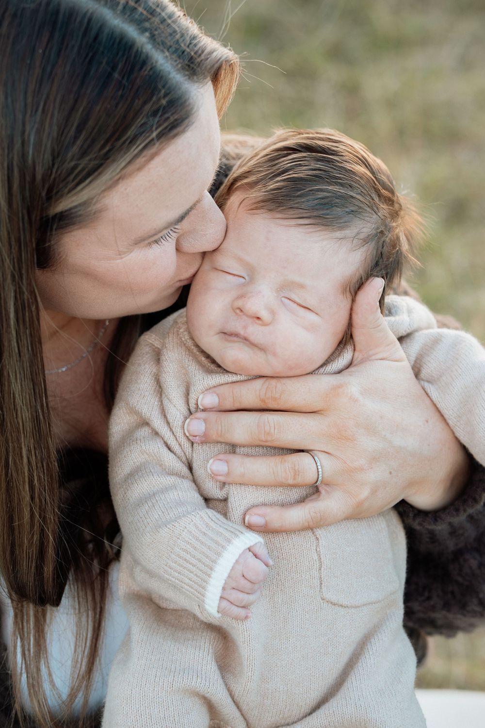 A mother tenderly kisses her sleeping newborn baby dressed in a beige onesie while cradling them gently outdoors.