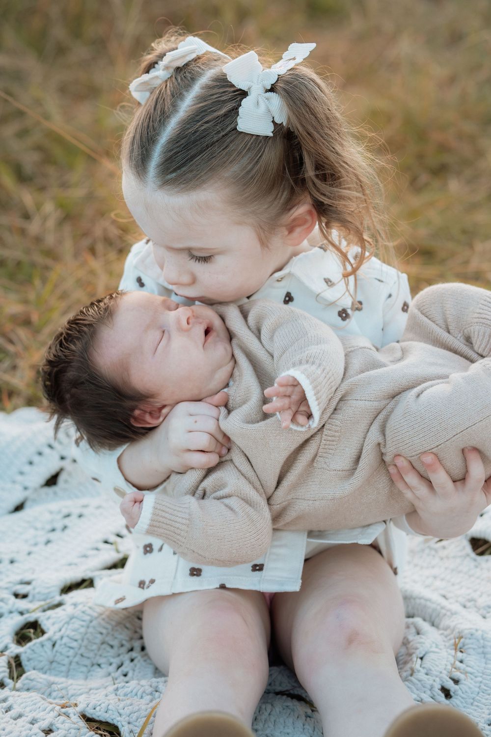 Young girl with bow pigtails kisses newborn baby sibling while sitting on crochet blanket outdoors.