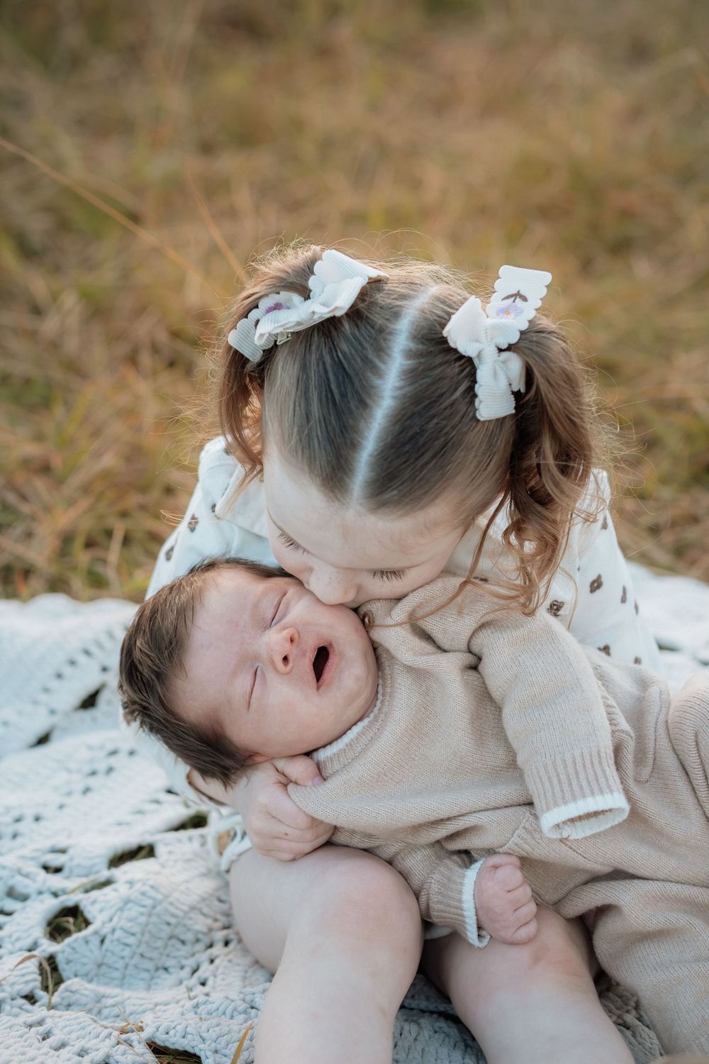 Young girl with pigtail bows kisses crying newborn baby sibling on a white blanket outdoors in Rouse Hill.