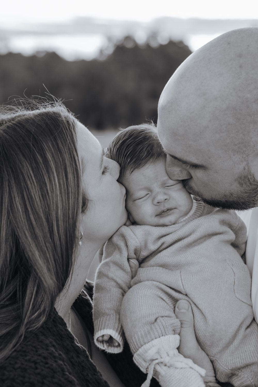 Black and white photo of parents kissing their newborn baby on the cheeks outdoors.