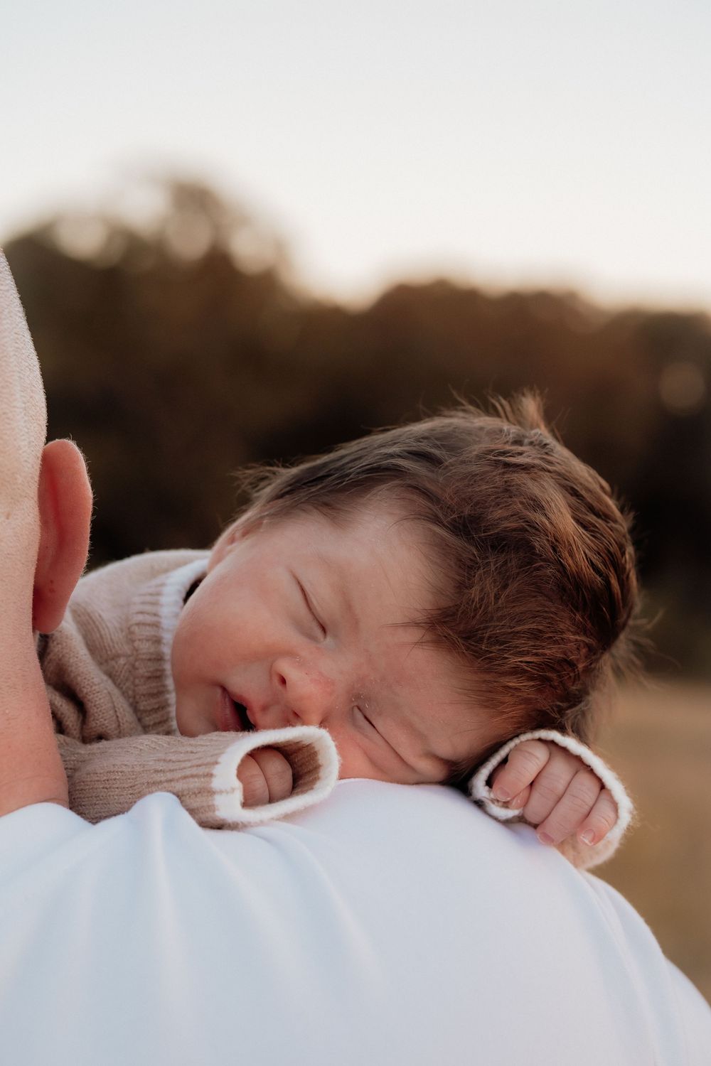 Peaceful newborn baby sleeping on fathers shoulder wearing beige knit romper in golden light.