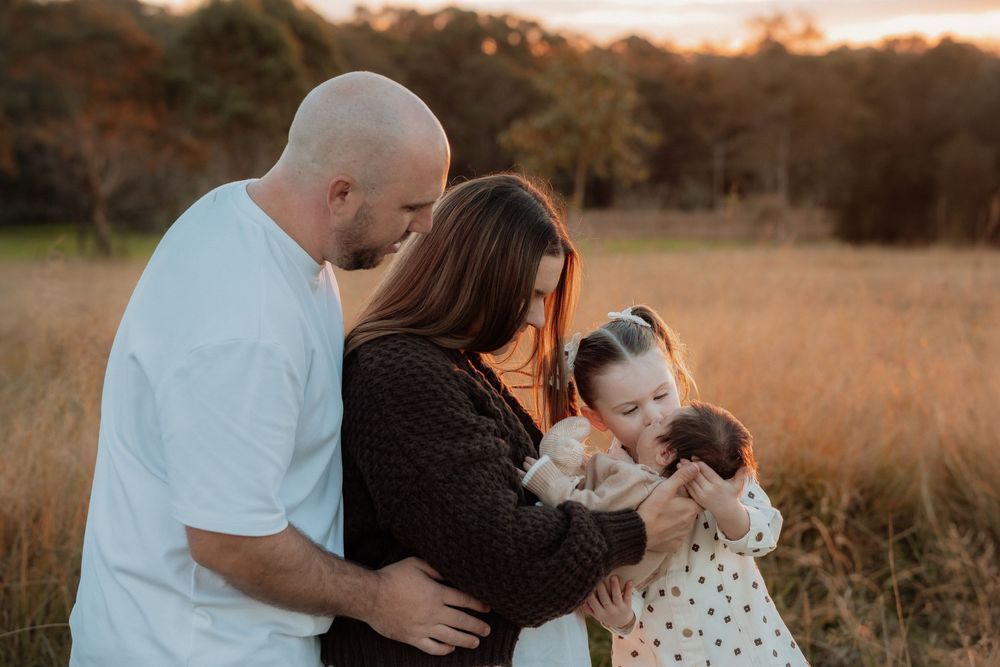 Family of four shares tender moment in golden field in Rouse HIll, toddler girl kissing newborn sibling.