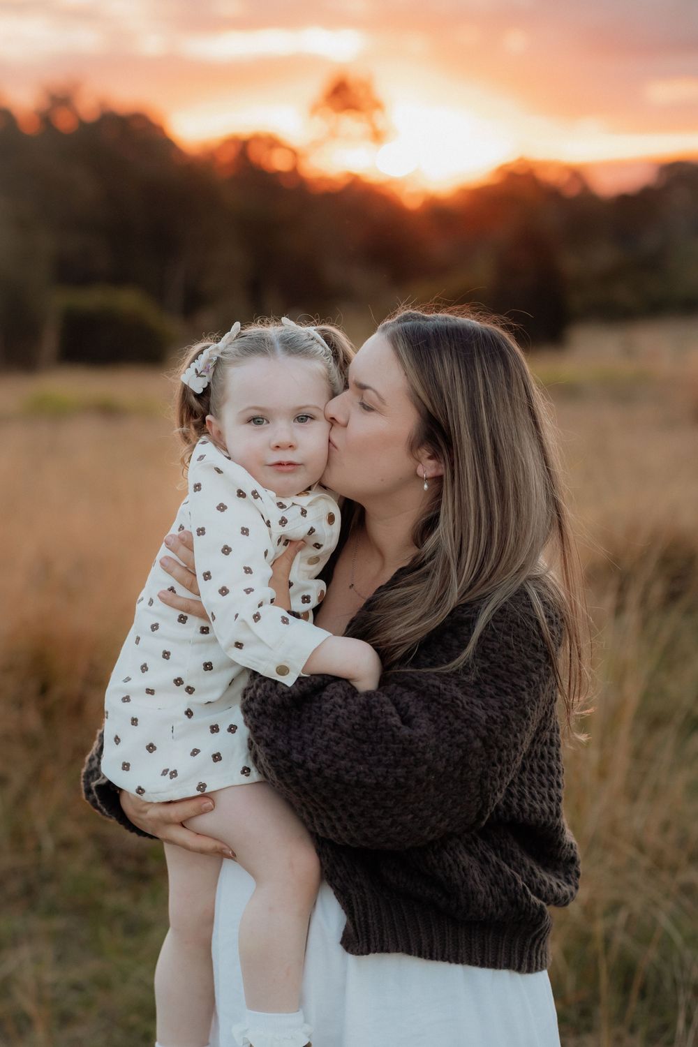 A mother tenderly kisses her toddler daughter on the cheek during a golden sunset outdoors at Rouse Hill Regional Park.