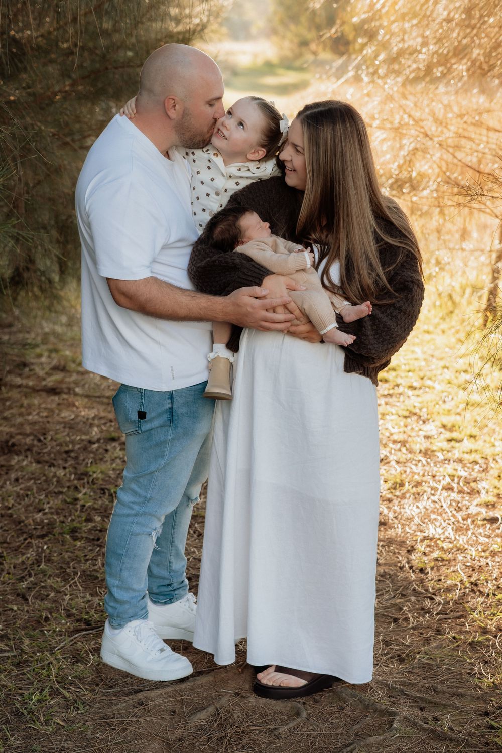 Family of four poses outdoors, parents holding toddler and newborn baby on a sunlit forest path at Rouse Hill Regional Park.