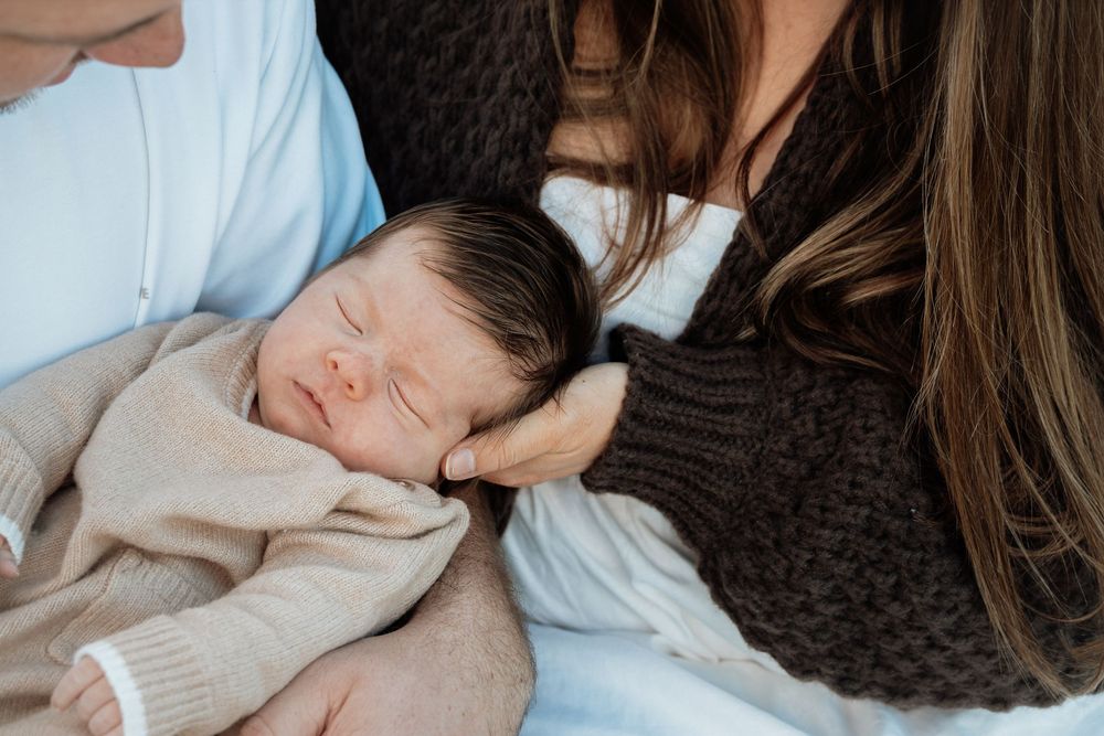 Sleeping newborn baby cradled in parents arms, wrapped in a soft beige blanket during a family portrait session.
