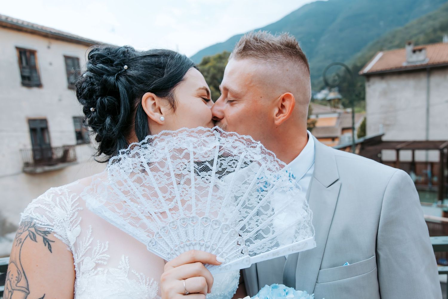 Bride and groom sharing a romantic kiss behind a white lace fan on their wedding day with mountains in background.